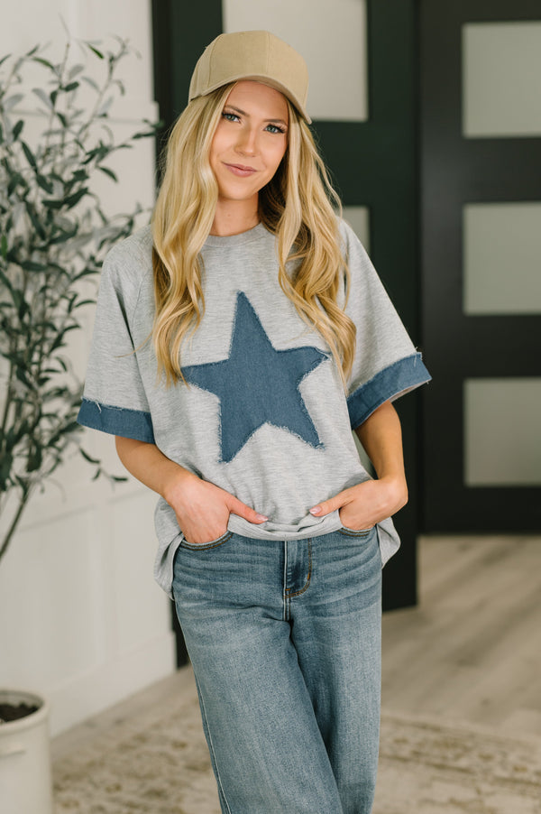 Woman wearing a gray sweater with a blue star, blue jeans, and a beige cap indoors.
