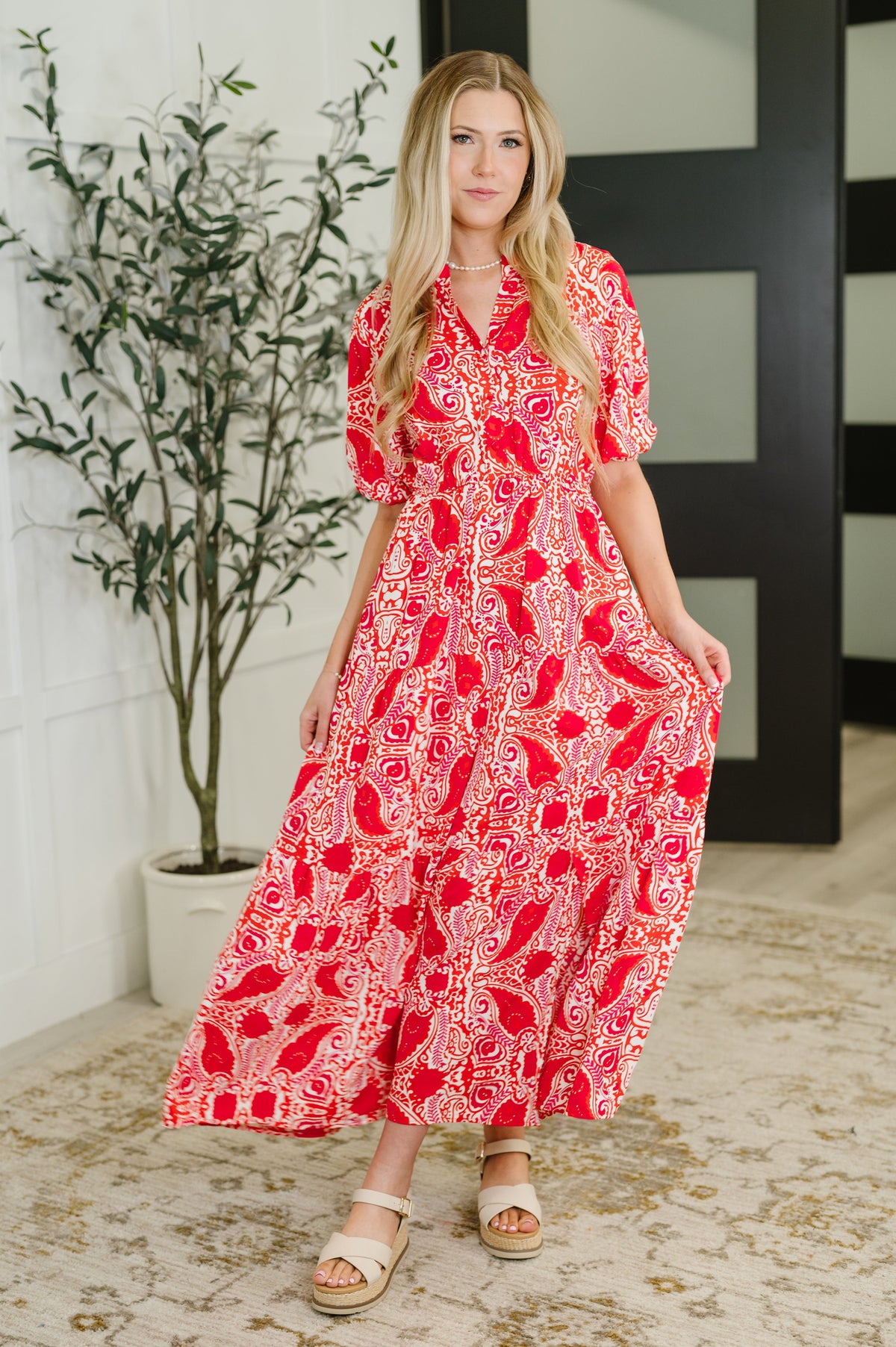 Woman wearing a red and white patterned dress standing indoors with a plant in the background.