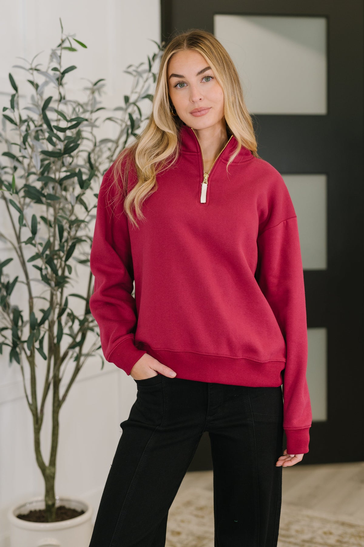 Woman wearing a red pullover with a white wall and plant in the background