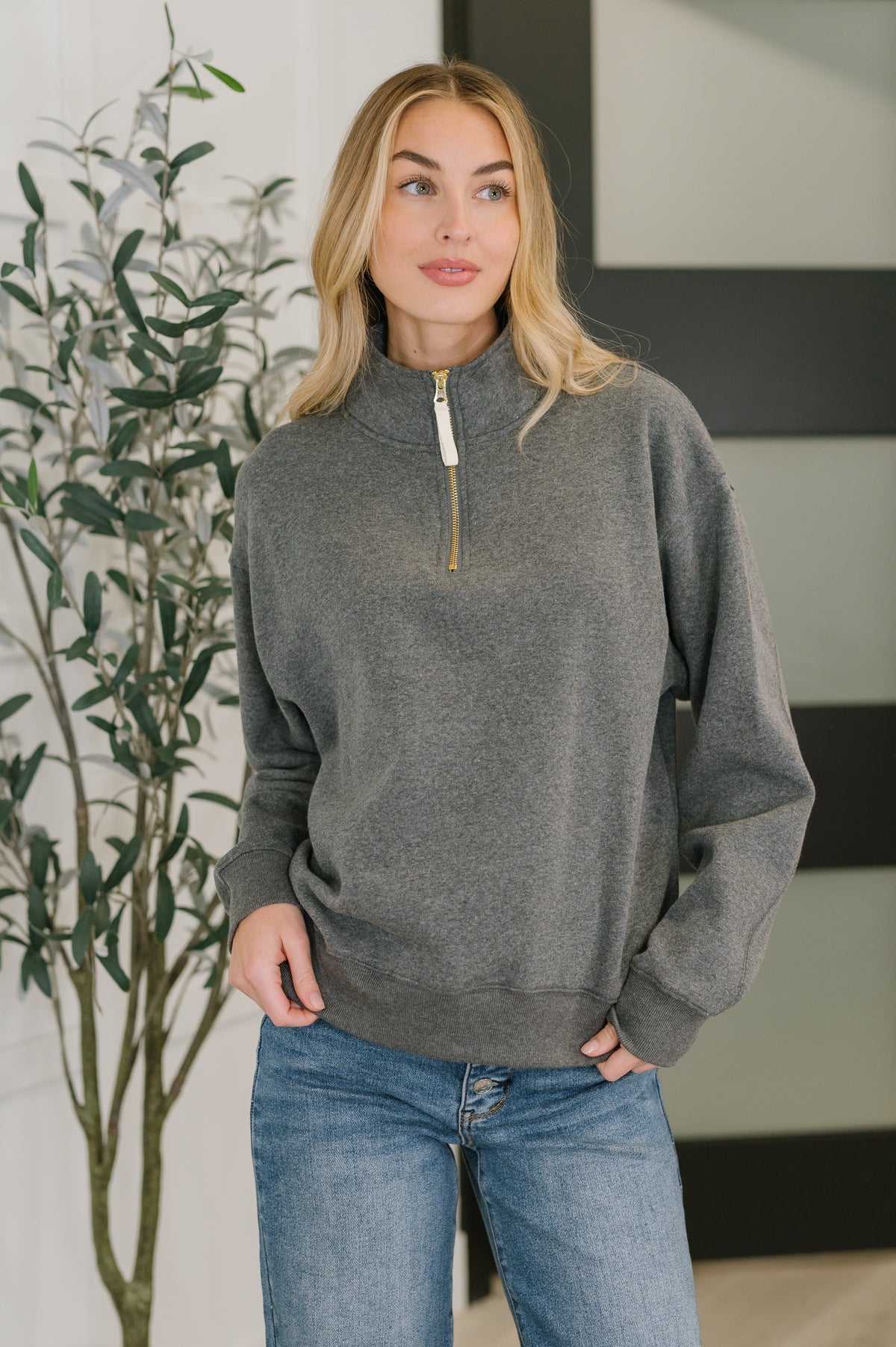 Woman wearing a gray sweater and blue jeans standing indoors with a plant in the background