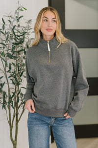 Woman wearing a gray sweater and blue jeans standing indoors with a plant in the background