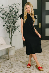 Woman wearing a black dress standing in a room with a plant and door in the background
