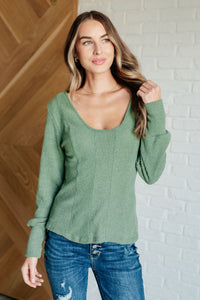 Woman wearing a green sweater and blue jeans against a white brick wall.