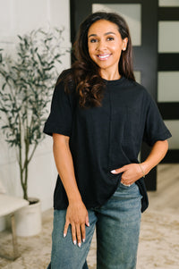 A woman wearing a black short-sleeve top and blue jeans, standing in a modern indoor setting with a potted plant and a black-and-white paneled wall in the background.