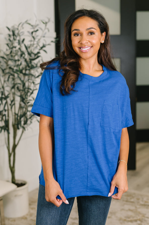 A woman wearing a short-sleeve, crew-neck blue top and dark wash jeans, standing in an indoor setting with a potted plant in the background.