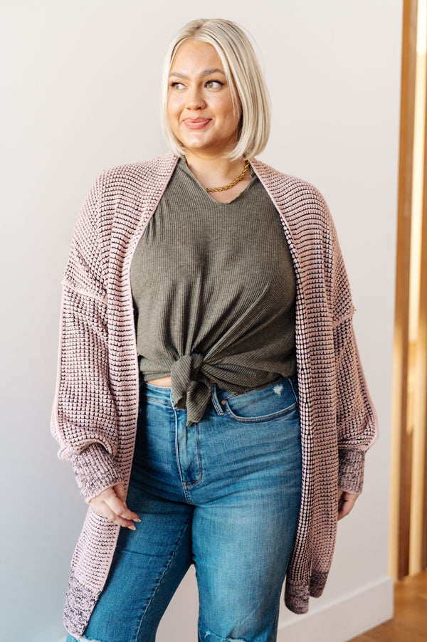 Woman wearing a pink cardigan over a green shirt and blue jeans against a white wall.