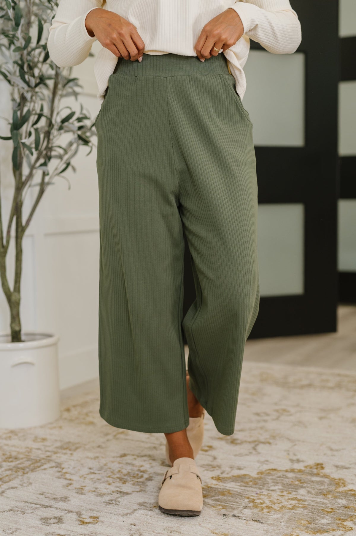 Person wearing green pants and beige shoes in a room with a plant and decorative wall.