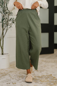 Person wearing green pants and beige shoes in a room with a plant and decorative wall.