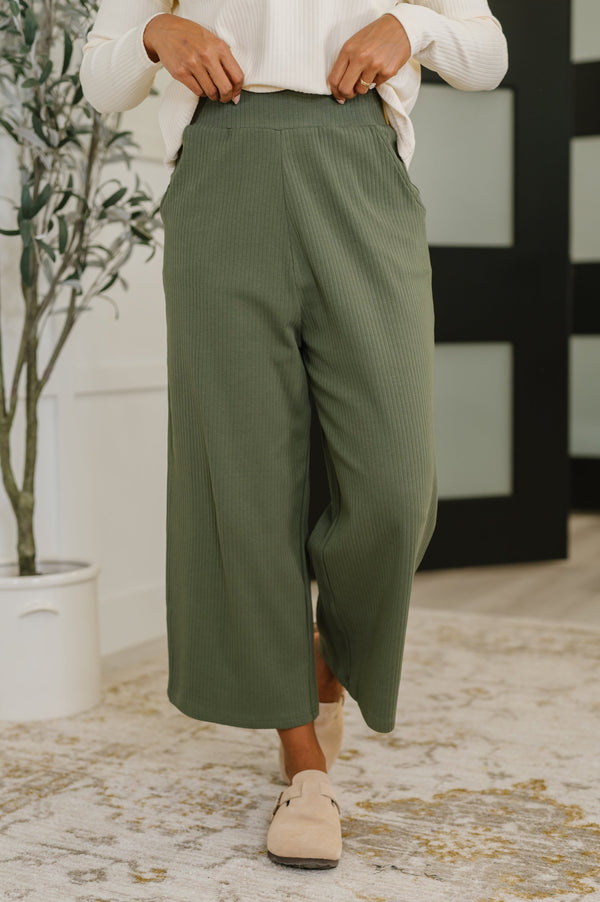Person wearing green pants and beige shoes in a room with a plant and decorative wall.