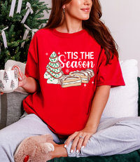 Person wearing a red 'Tis the Season t-shirt with Christmas tree and cookies design, holding a mug.
