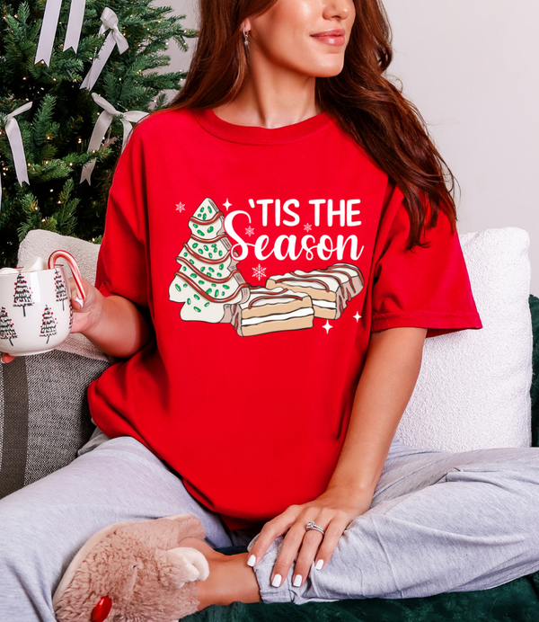 Person wearing a red 'Tis the Season t-shirt with Christmas tree and cookies design, holding a mug.