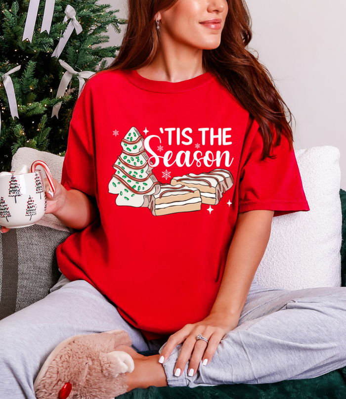 Person wearing a red 'Tis the Season t-shirt with Christmas tree and cookies design, holding a mug.