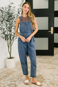 Woman wearing a denim jumpsuit standing indoors with a plant and door in the background