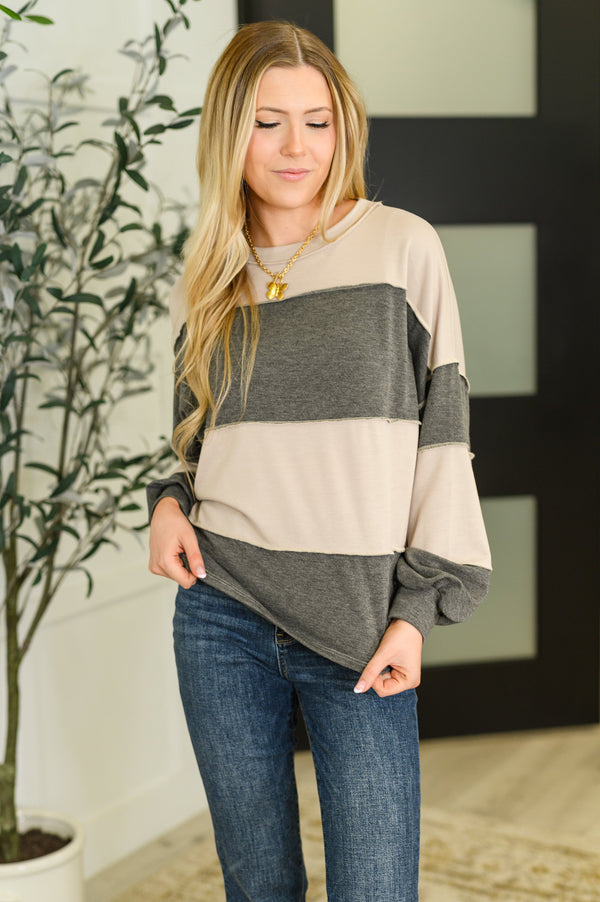 Woman wearing a beige and gray color-blocked sweater with blue jeans indoors.