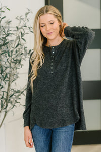 Woman wearing a dark gray long-sleeve shirt and blue jeans, standing indoors with a plant in the background.