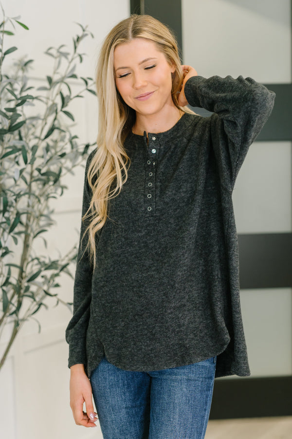 Woman wearing a dark gray long-sleeve shirt and blue jeans, standing indoors with a plant in the background.