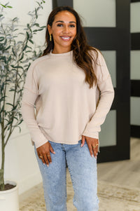 Woman wearing a beige sweater and blue jeans standing indoors with a plant in the background