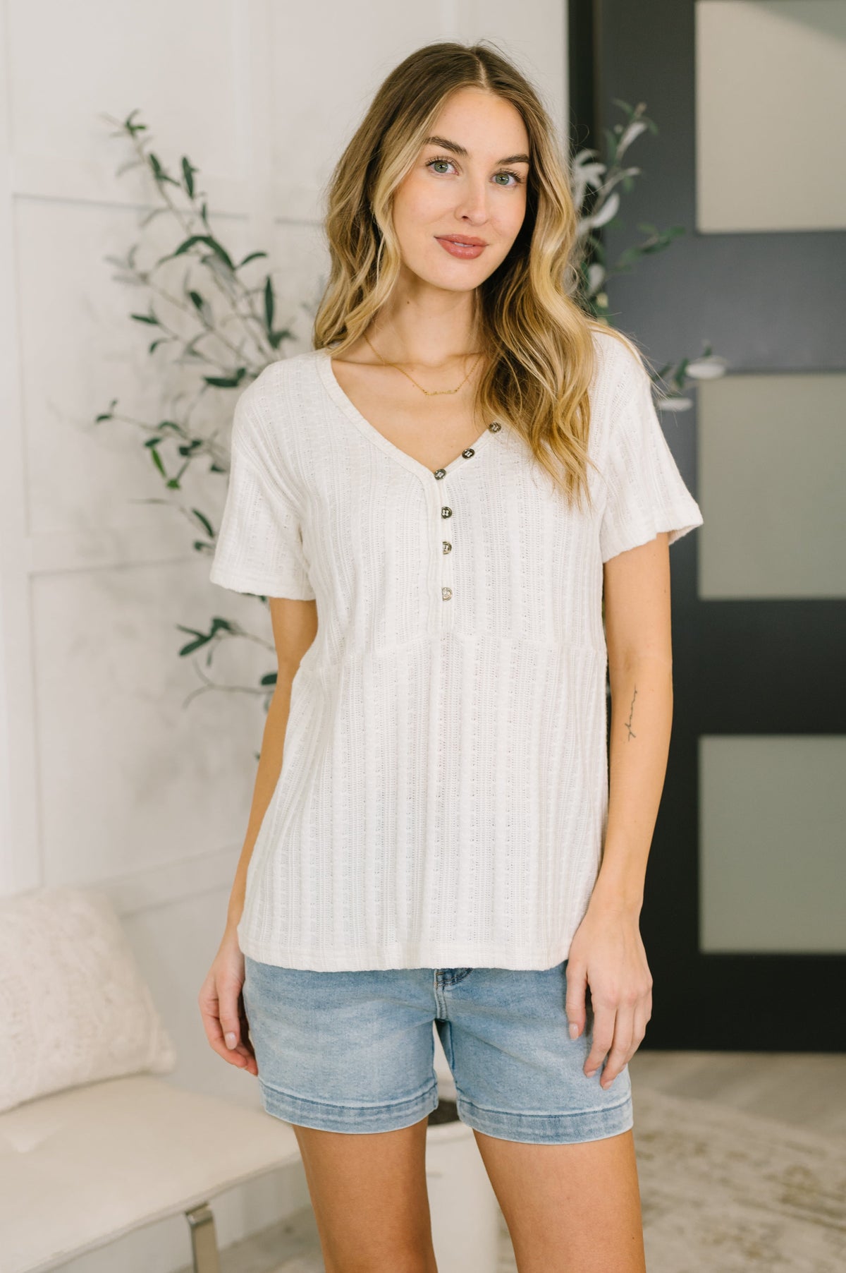 Woman wearing a white ribbed top and denim shorts in a room with decorative elements.