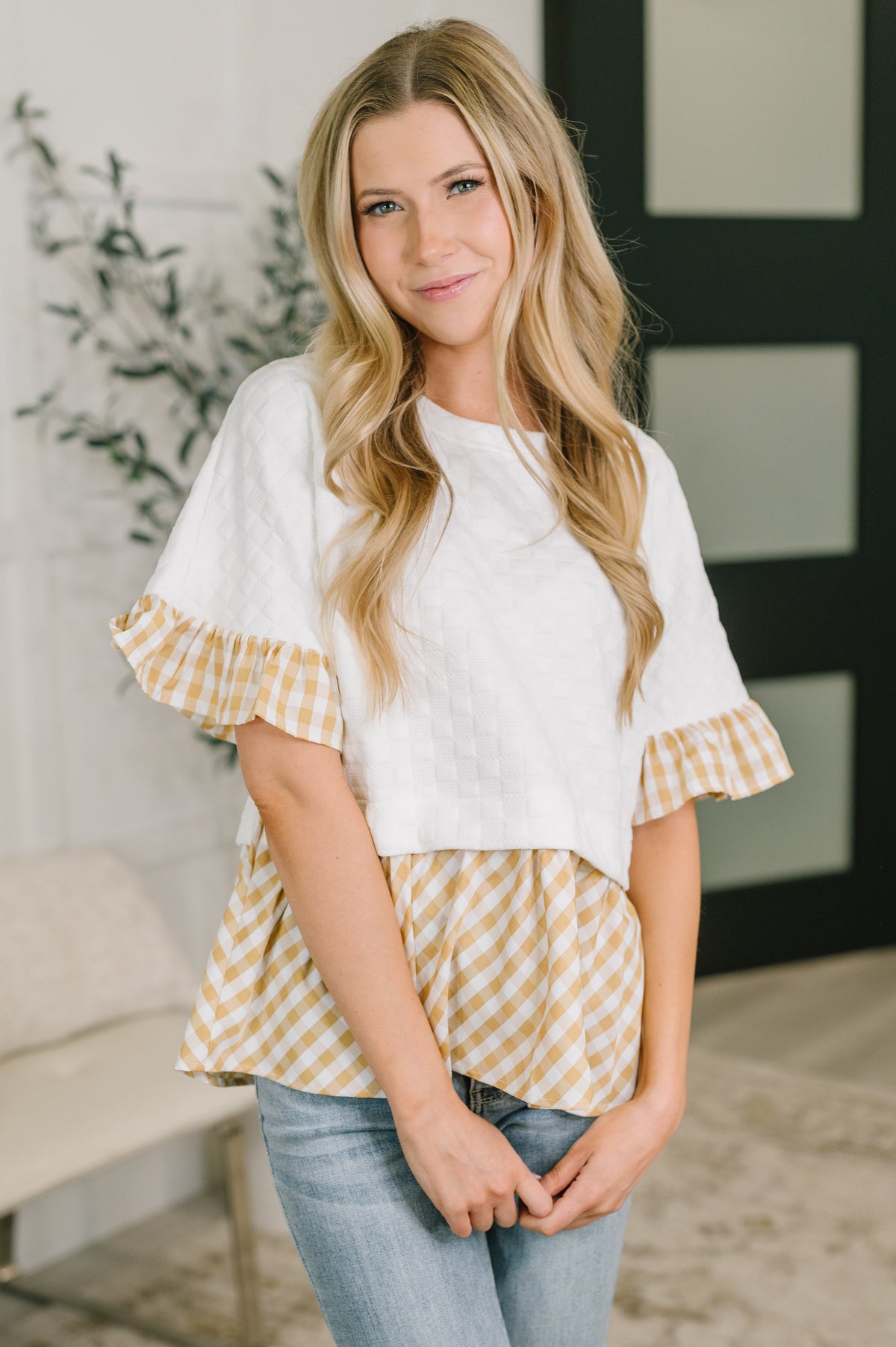 Woman wearing a white top with yellow plaid ruffle sleeves and blue jeans indoors.