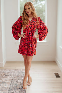A woman modeling a red and black leopard-print mini dress with dramatic balloon sleeves in a bright, modern room with light wood floors and a window behind her.