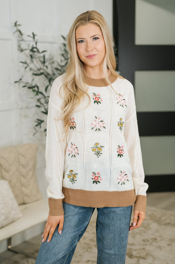 A woman modeling a cream-colored knit sweater with brown ribbed trim and pink, green, and yellow floral embroidery, paired with blue jeans in a modern indoor setting.