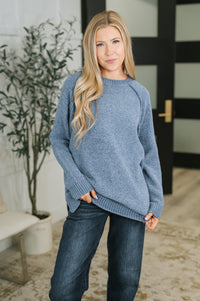 A woman wearing a heathered dusty blue knit sweater and dark blue jeans, standing in a brightly lit indoor setting with a potted plant and modern door in the background.