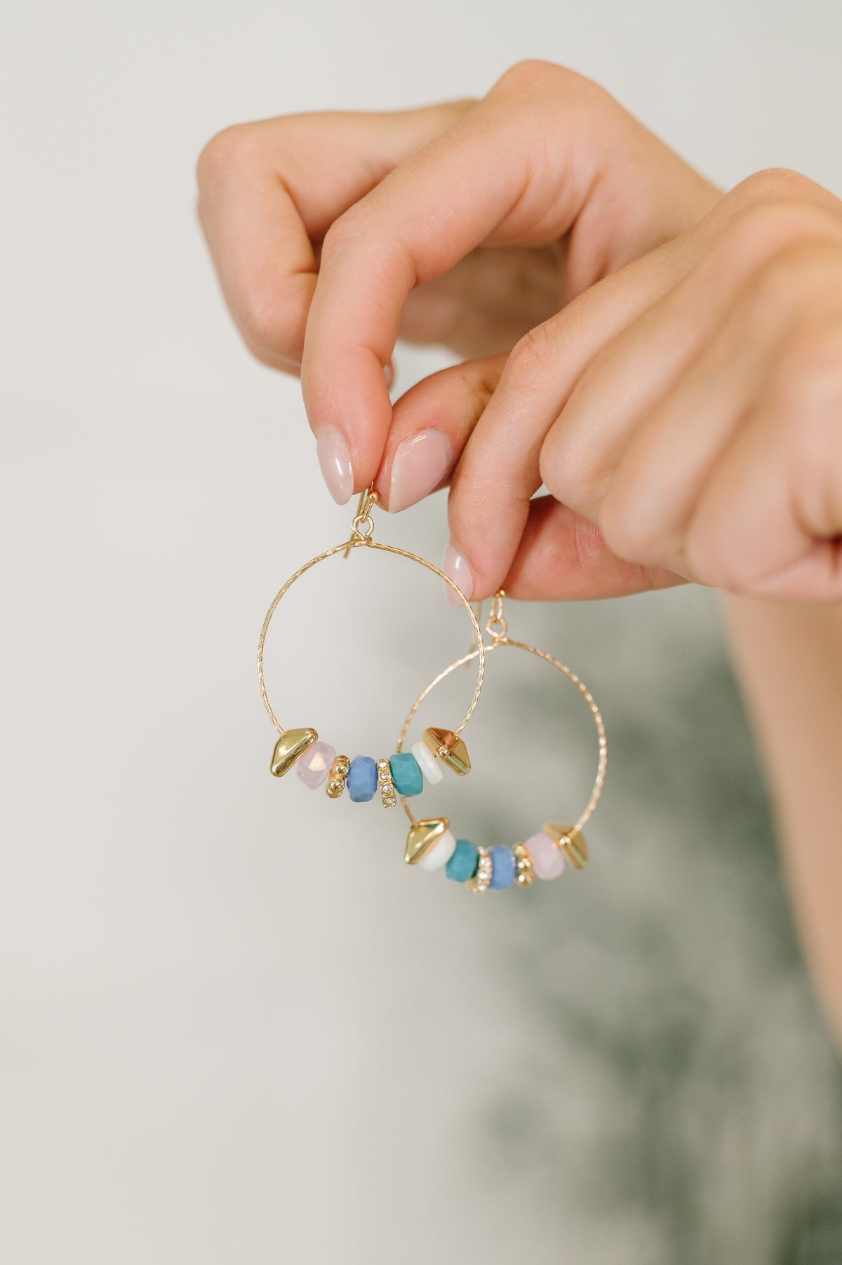 Gold hoop earrings with colorful beads held by a hand against a neutral background