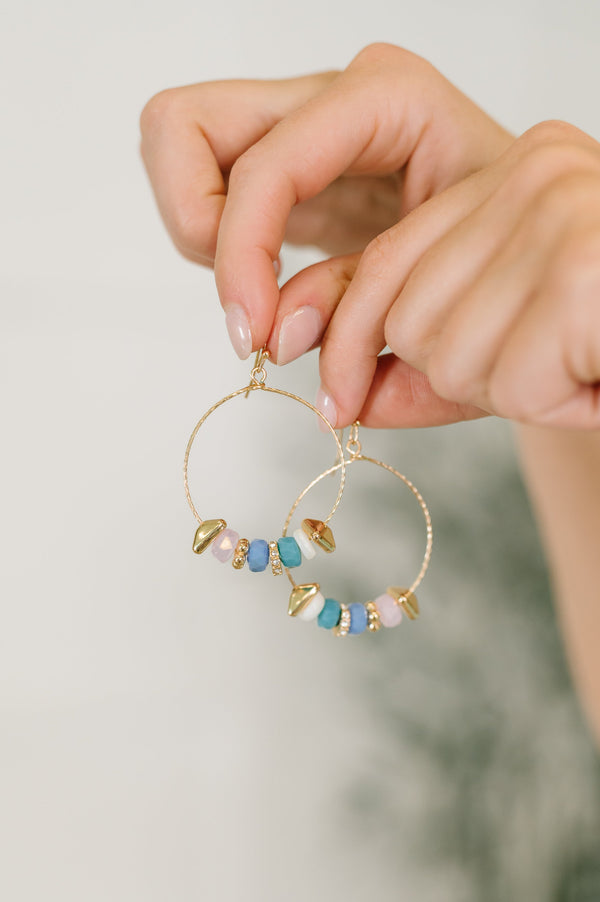Gold hoop earrings with colorful beads held by a hand against a neutral background