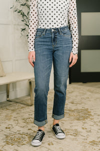 A person wearing a pair of medium-wash blue straight-leg jeans with rolled cuffs, paired with a white long-sleeve top with black polka dots and black low-top sneakers, standing on a patterned beige rug.