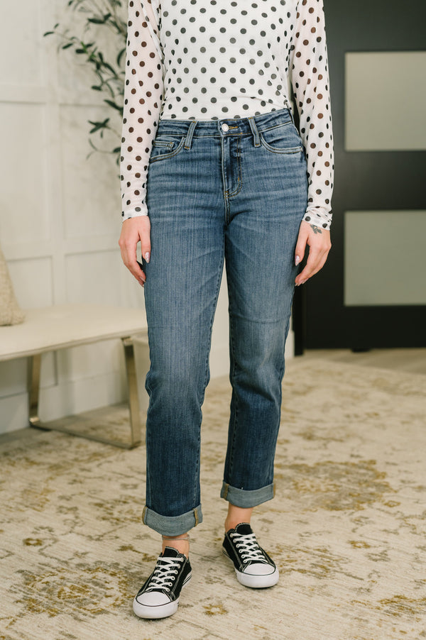 A person wearing a pair of medium-wash blue straight-leg jeans with rolled cuffs, paired with a white long-sleeve top with black polka dots and black low-top sneakers, standing on a patterned beige rug.