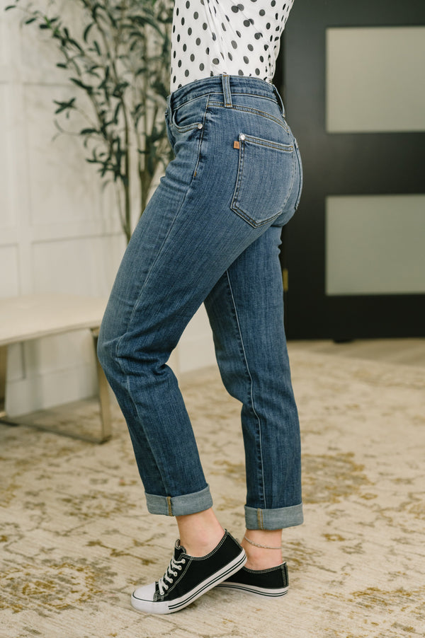 A person wearing blue cuffed jeans, a white top with black polka dots, and black low-top sneakers, shown from the waist down in an indoor setting.